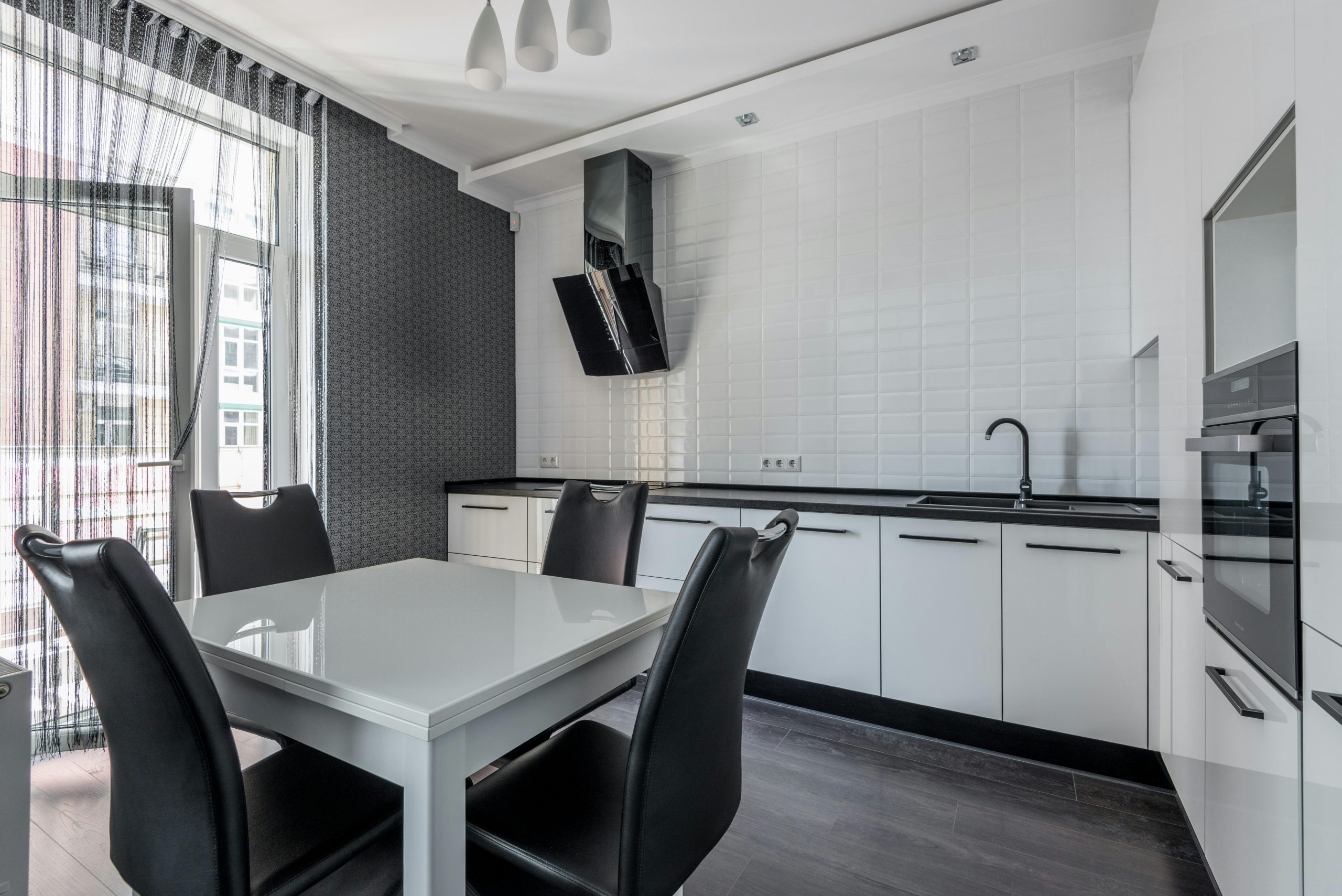 Stylish white kitchen furnished with white cupboards and built in oven near white table with big window